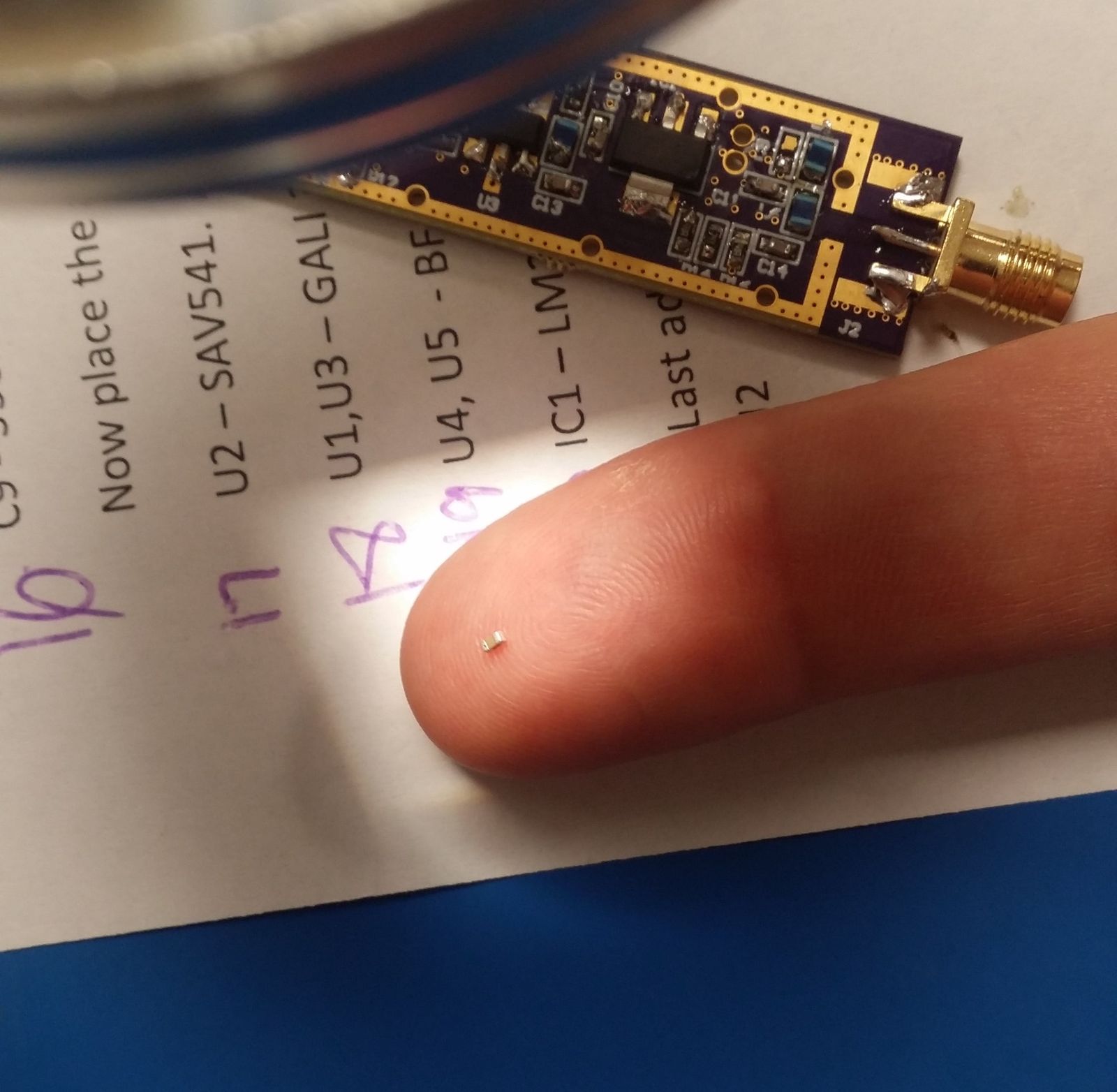 A surface-mount amplifier component a couple of millimeters across, balanced on a fingertip above a printed assembly sheet