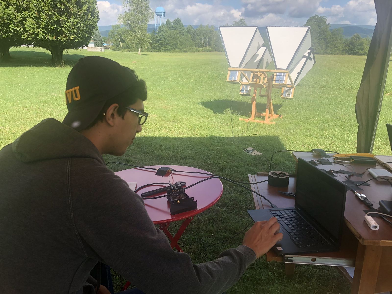 An undergraduate researcher working at a laptop outdoors, a horn aperture array set up in the field behind him