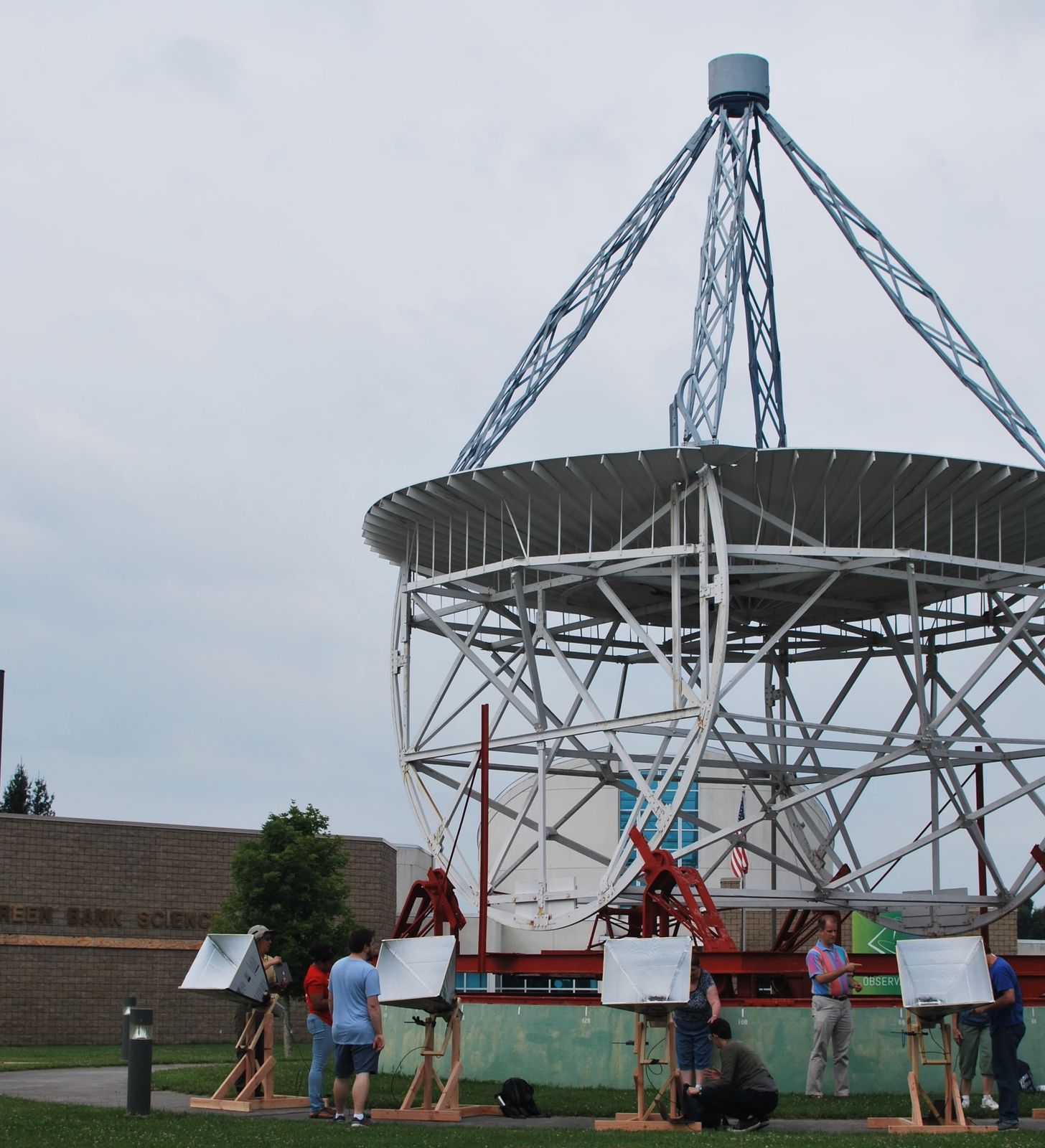 Four horn antennas on wooden mounts lined up on the grass outside the Green Bank Science Center, with the Reber Telescope towering behind them