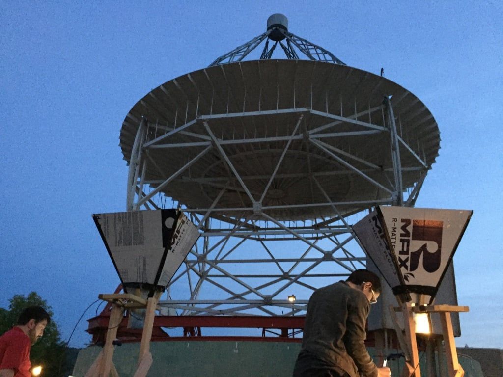 Two teachers working by lamplight with horn antennas beneath the Reber Telescope against a deep blue dusk sky